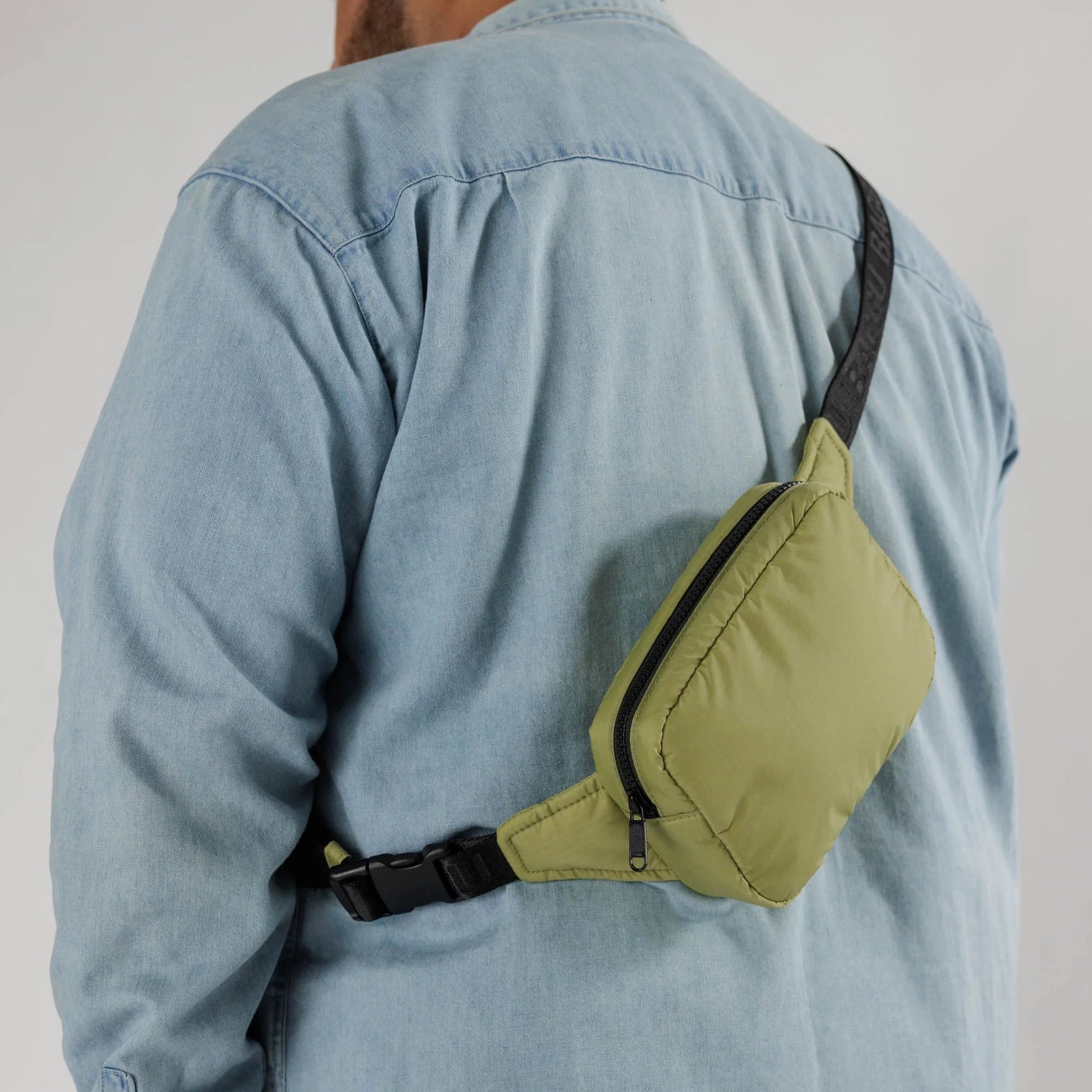 On a white background is the puffy fanny pack in the shade pistachio, which is an olive green color with a black adjustable strap that has subtle letters that read, &quot;BAGGU&quot; in all caps worn here on a model. 