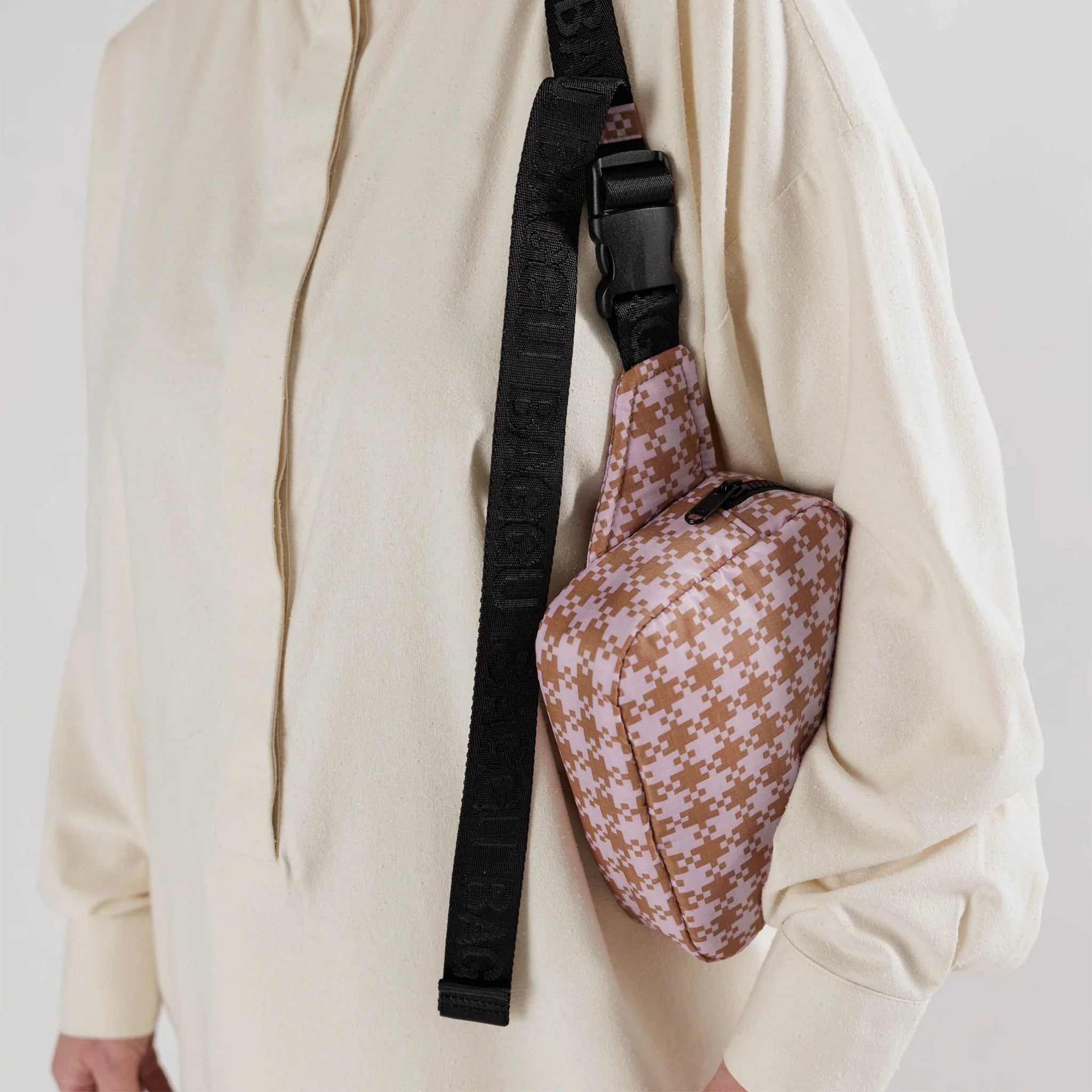 A model wearing a fanny pack with a brown and pink gingham pattern and a black adjustable strap. 