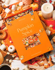 An orange puzzle box that reads "Pumpkin Spice" site on top of a half-worked puuzzle below. In the backround are lit white candles, popcorn sprinkled about, marshmallows sprinkled about, an old fashioned donut and a cup of cofee. The image evokes the feeling of Fall colors and mood.