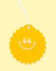On a yellow background is a yellow sunshine shaped air freshener with a smiley face and a white hanging string.