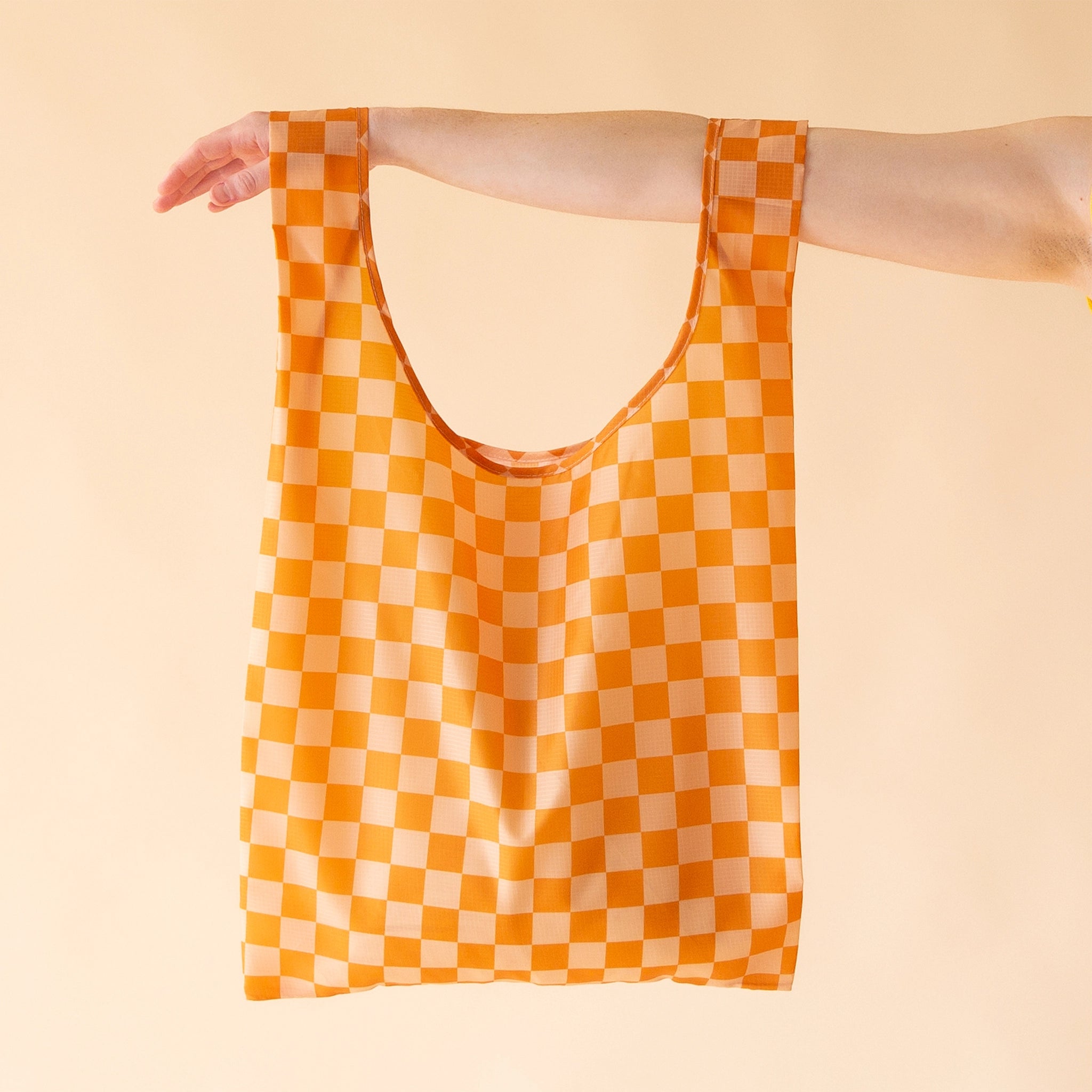 On a cream background is the Playa Check Reusable Bag which features two shoulder handles and a dark orange and lighter orange checker print. 