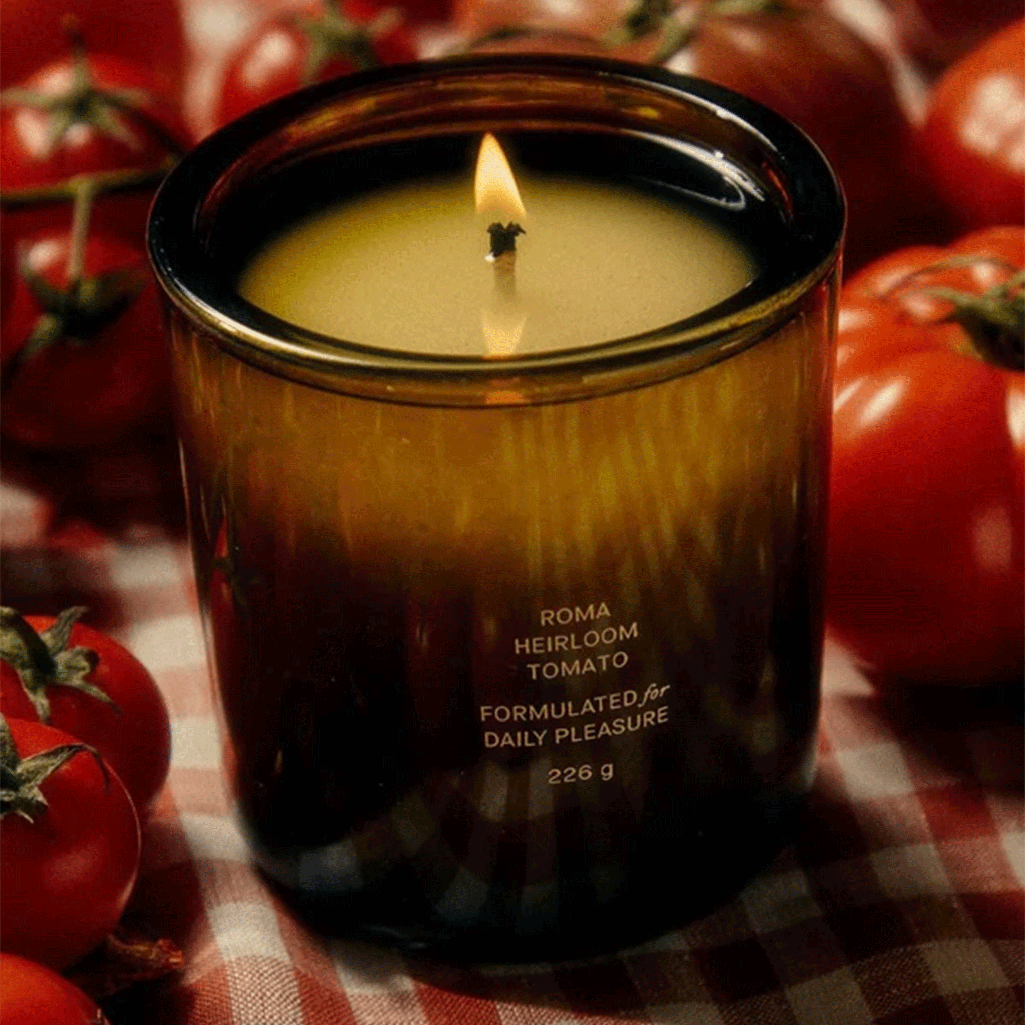 A green glass candle with small text in the front that reads, "Roma Heirloom Tomato Formulated for Daily Pleasure" surrounded by red tomatoes. 