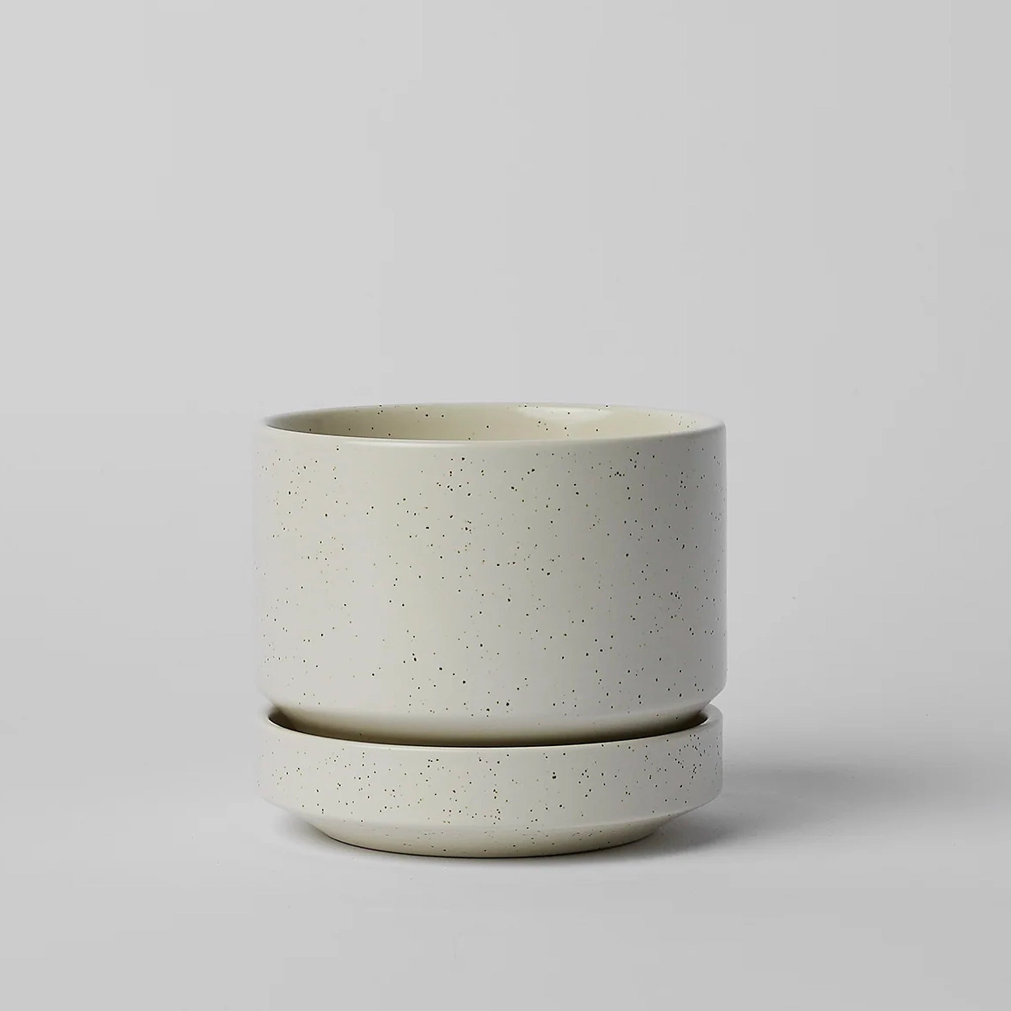 On a neutral background is a cream ceramic planter with tiny black speckles and a removable tray for watering. 