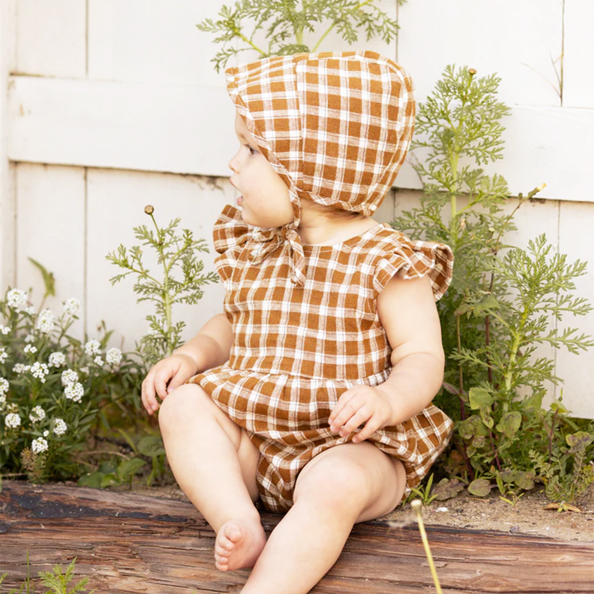 A model wearing a brown and ivory plaid one piece romper with ruffle detials on the sleeves. Bonnet not included.