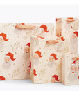 On a tan background is three different sized ivory gift bags with a red Santa design along with gold text that reads, "Merry Christmas".