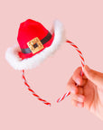 Hand holding a small Santa hat headband with candy canes detail on the headband against a pink background