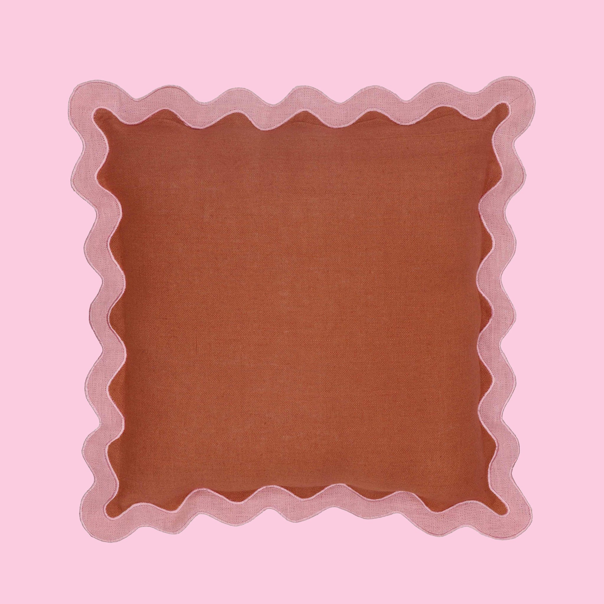 On a pink background is a scalloped edge throw pillow with a terracotta center and a wavy pink edge. 