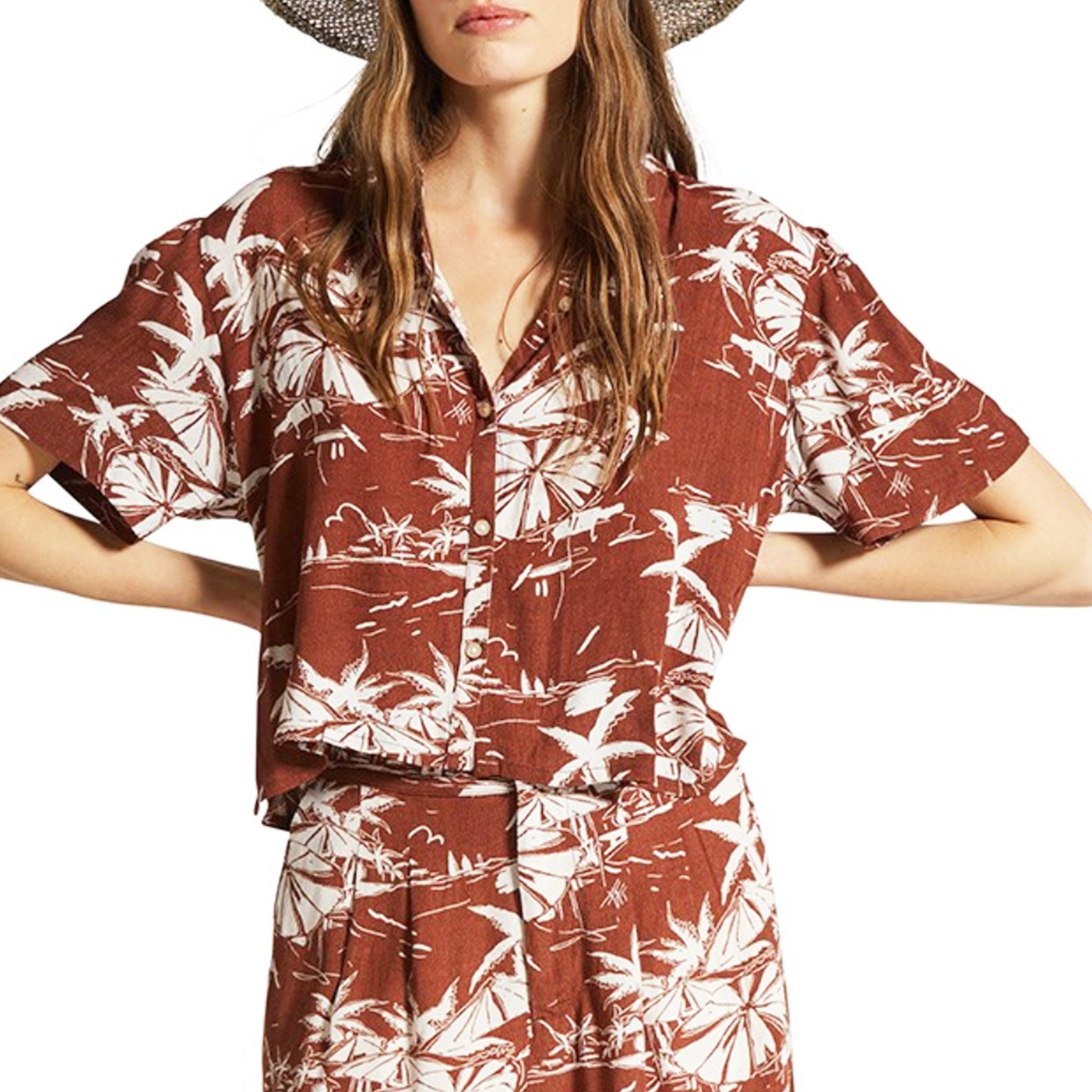 A rust colored button up short sleeve shirt with an ivory tropical print. 