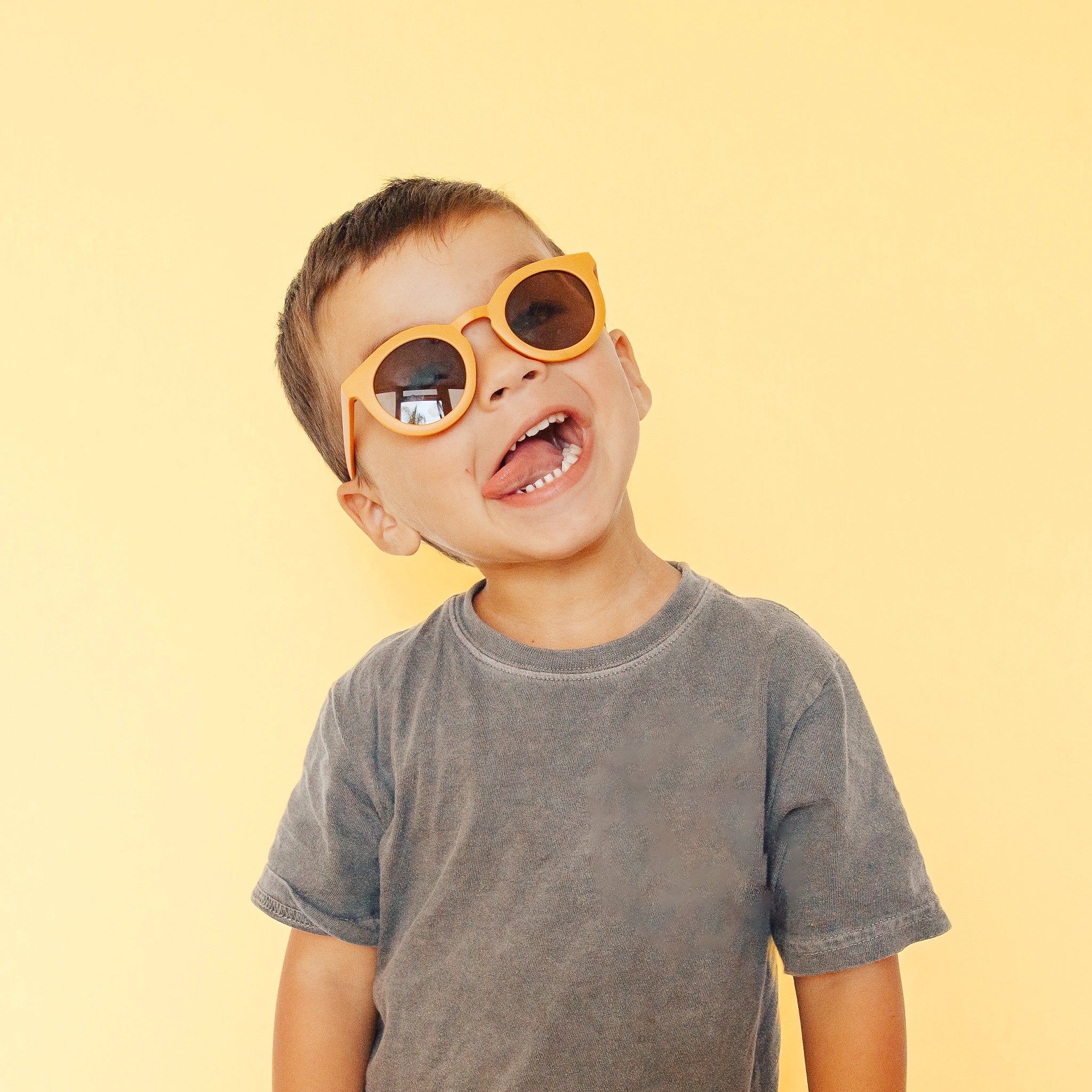 On a light yellow background is a children&#39;s model wearing a yellow round pair of sunglasses. 