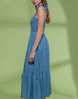 On a green shadow background is a model wearing a blue flowy midi dress with tie shoulder straps and a square neckline.