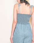 A light blue denim smocked jumpsuit with tie shoulder straps.