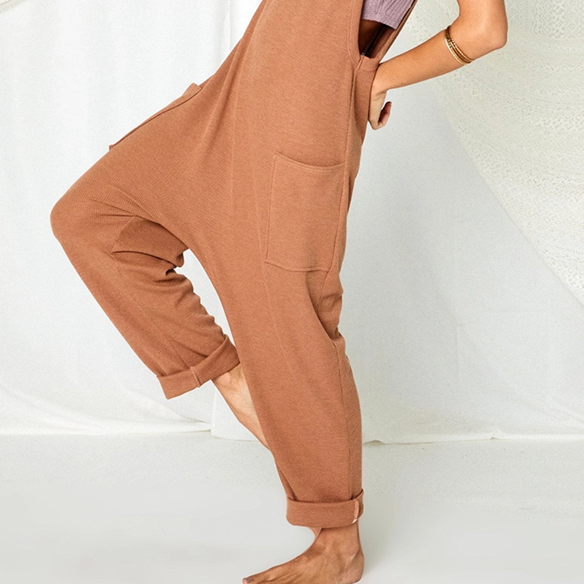 On a white background is a model wearing a camel colored jumpsuit with a drop crotch, front pockets and a v-neck.