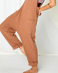 On a white background is a model wearing a camel colored jumpsuit with a drop crotch, front pockets and a v-neck.