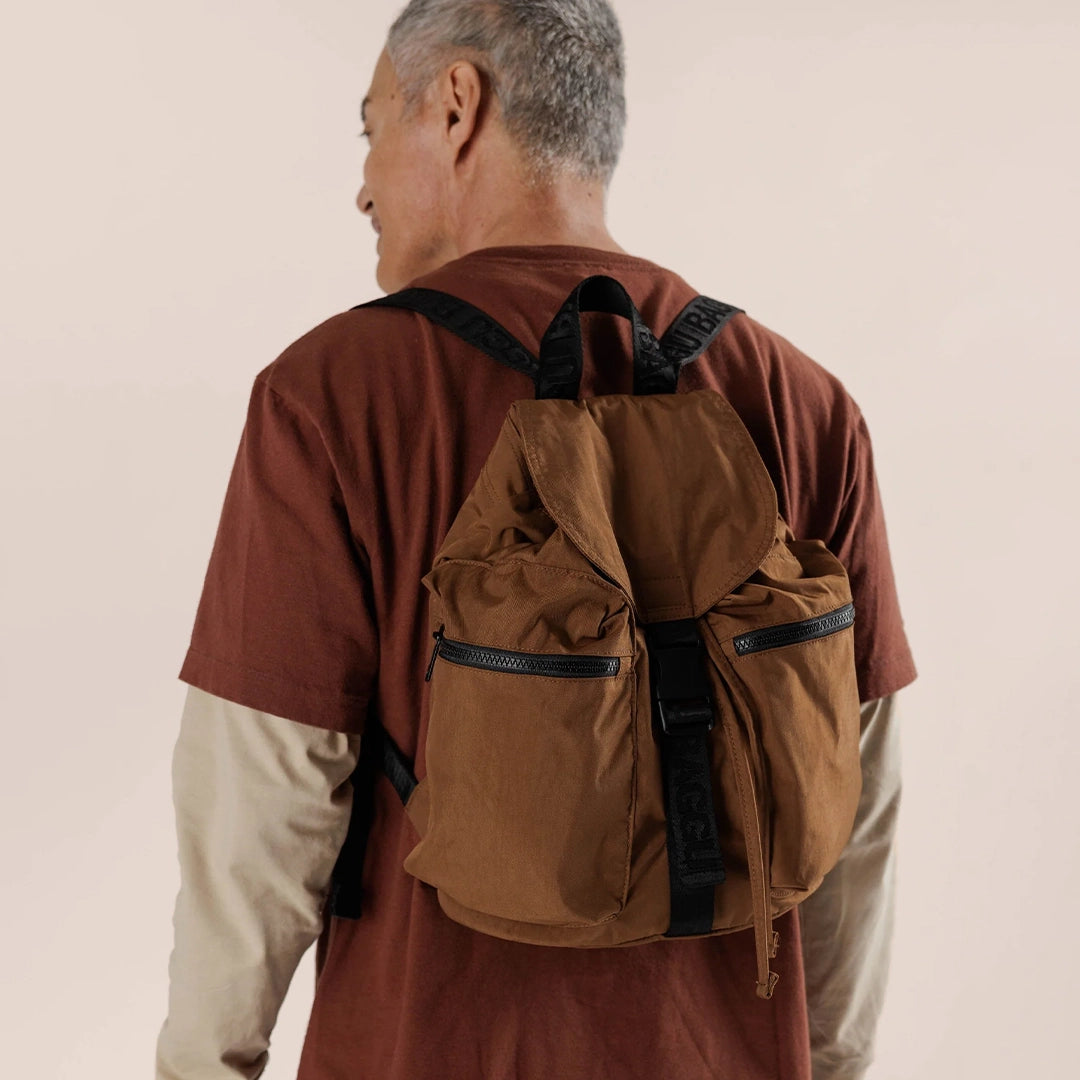 On a cream background is a brown nylon backpack with a drawstring closure with a flap overtop that buckles in the front as well as black straps and two side pockets.