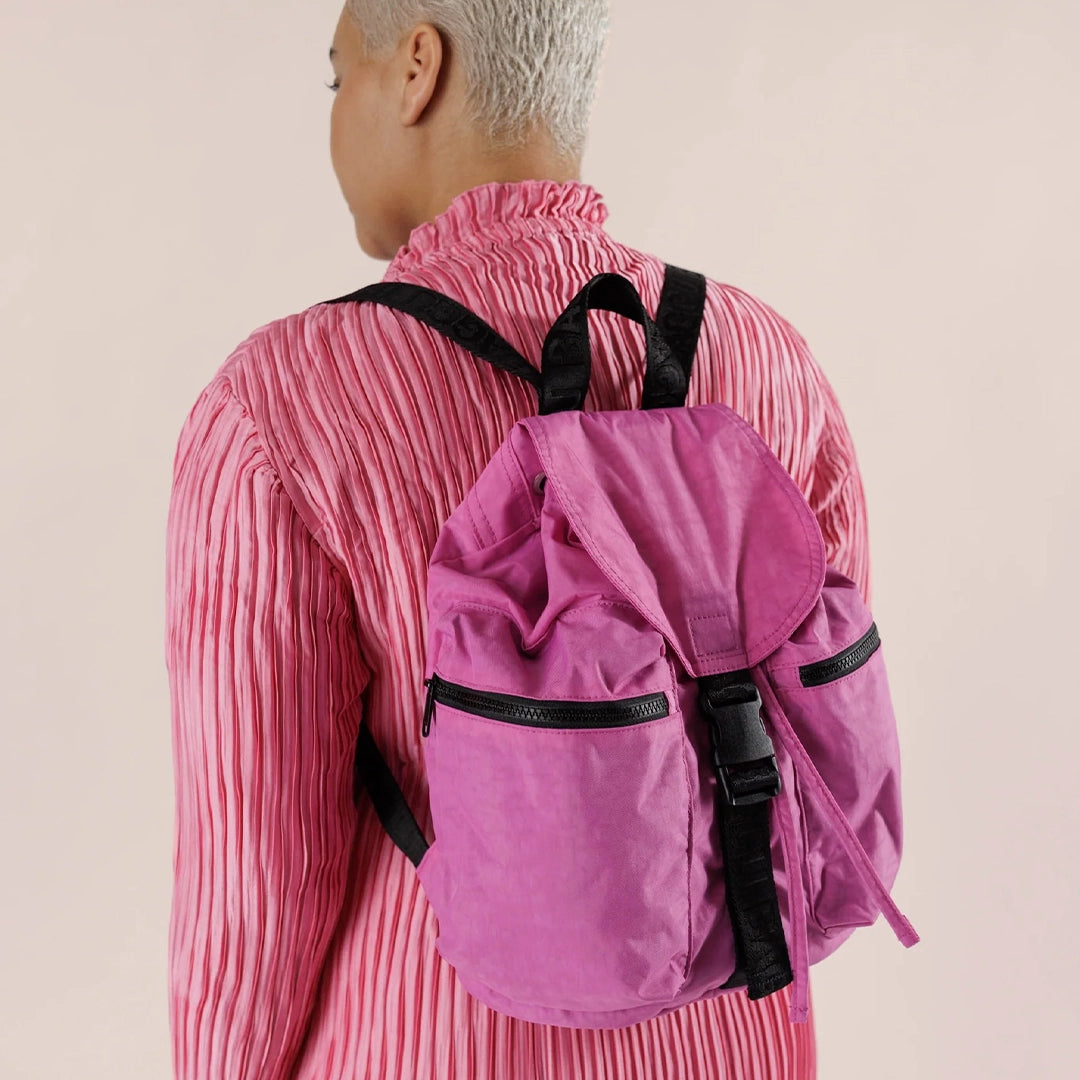 On a cream background is a bright pink nylon backpack with a drawstring closure with a flap overtop that buckles in the front as well as black straps and two side pockets.