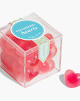 In front of a white background is the birds eye view of a clear, plastic cube. There is a thin, blue and gold sticker that moves vertically down the middle of the lid. In the middle is a round blue sticker with white text that reads ‘strawberry hearts.’ The cube is filled with bright pink gummies.