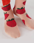 On a white background is a model wearing pink crew socks with a red strawberry pattern on it.