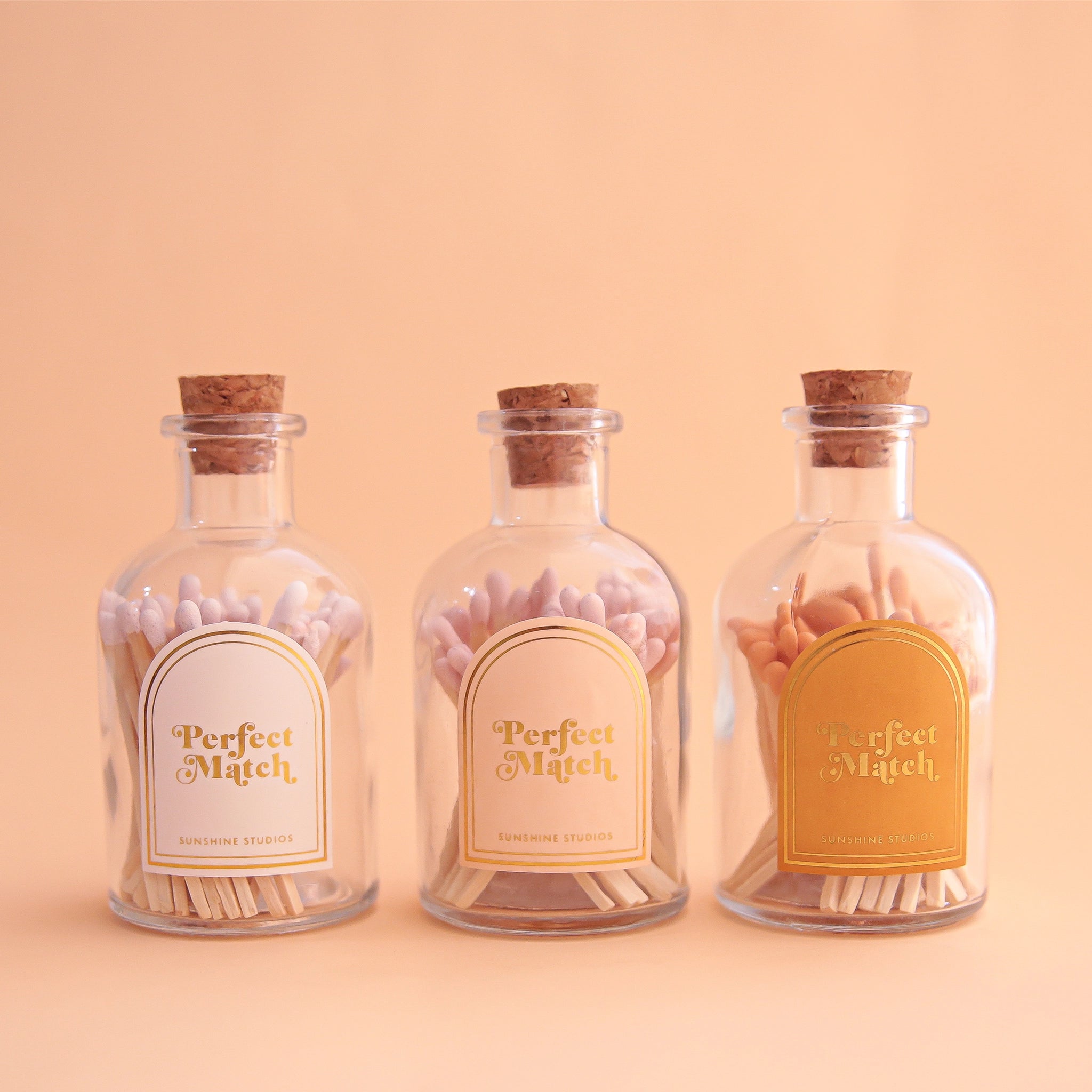 Three glass jars filled with wooden matches and a cork stopper. Each jar has an arched label that reads, "Perfect Match" in white, pink and gold. 