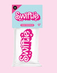A pink and white air freshener in the plastic packaging that reads, "Swiftie".