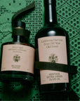 Two dark green Flamingo Estate bottles—one balsamic vinegar and one heritage extra virgin olive oil—rest on a textured green lace cloth, each with cream labels and green ribbon accents.
