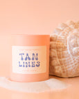 An apricot colored glass candle with a tan label, blue text that reads, 'Tan Lines'.