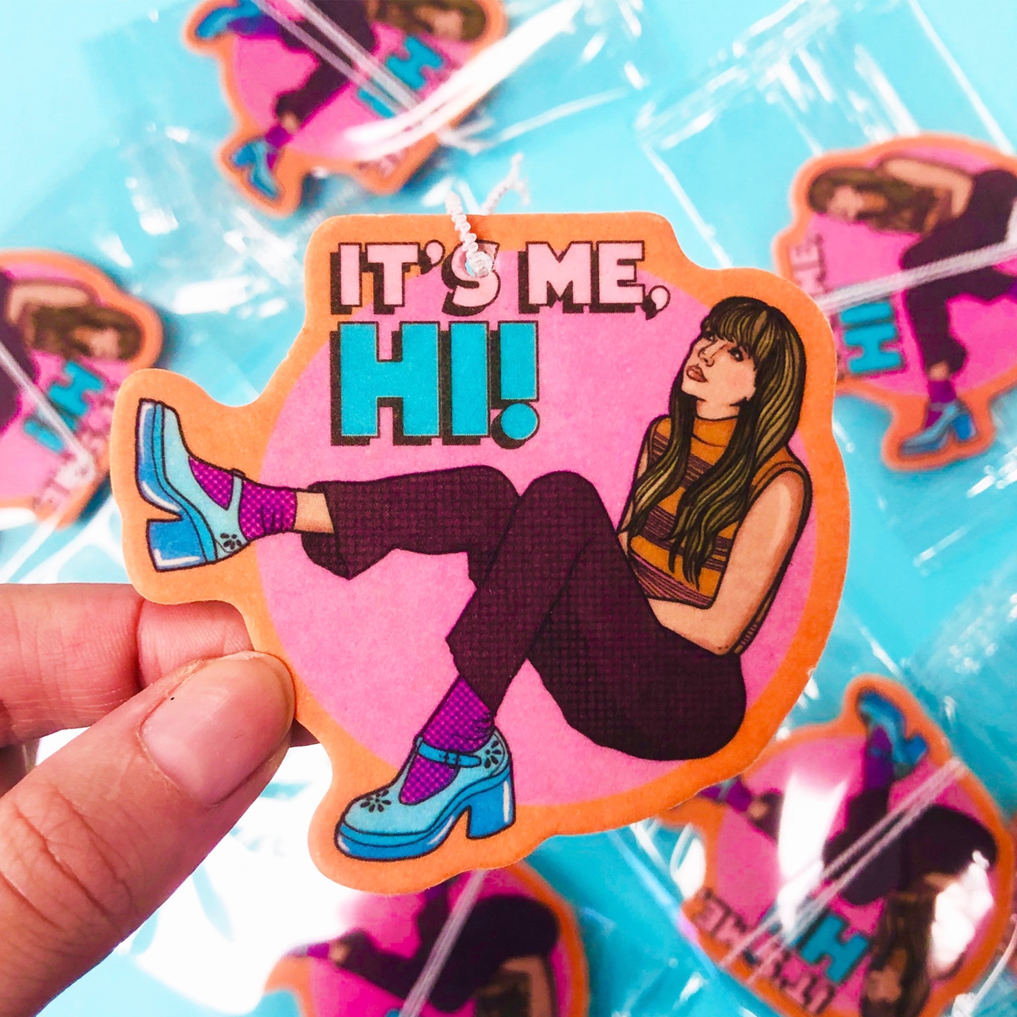 An air freshener with a graphic that looks like Taylor Swift with text that reads, "It's Me, Hi!". 