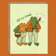 Card with two frogs in autumn-themed clothing and accessories on a beige background.
