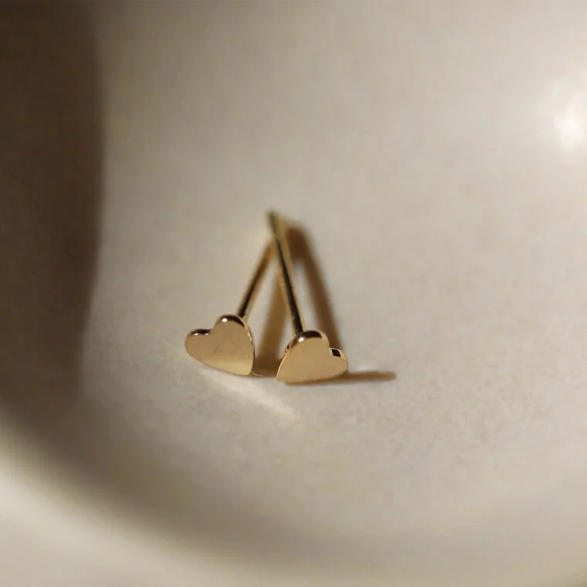 In a cream ceramic bowl is a pair of gold stud earrings in the shape of tiny hearts. 