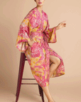 On a tan background is a model wearing a pink and yellow/orange robe with a tropical and floral print.