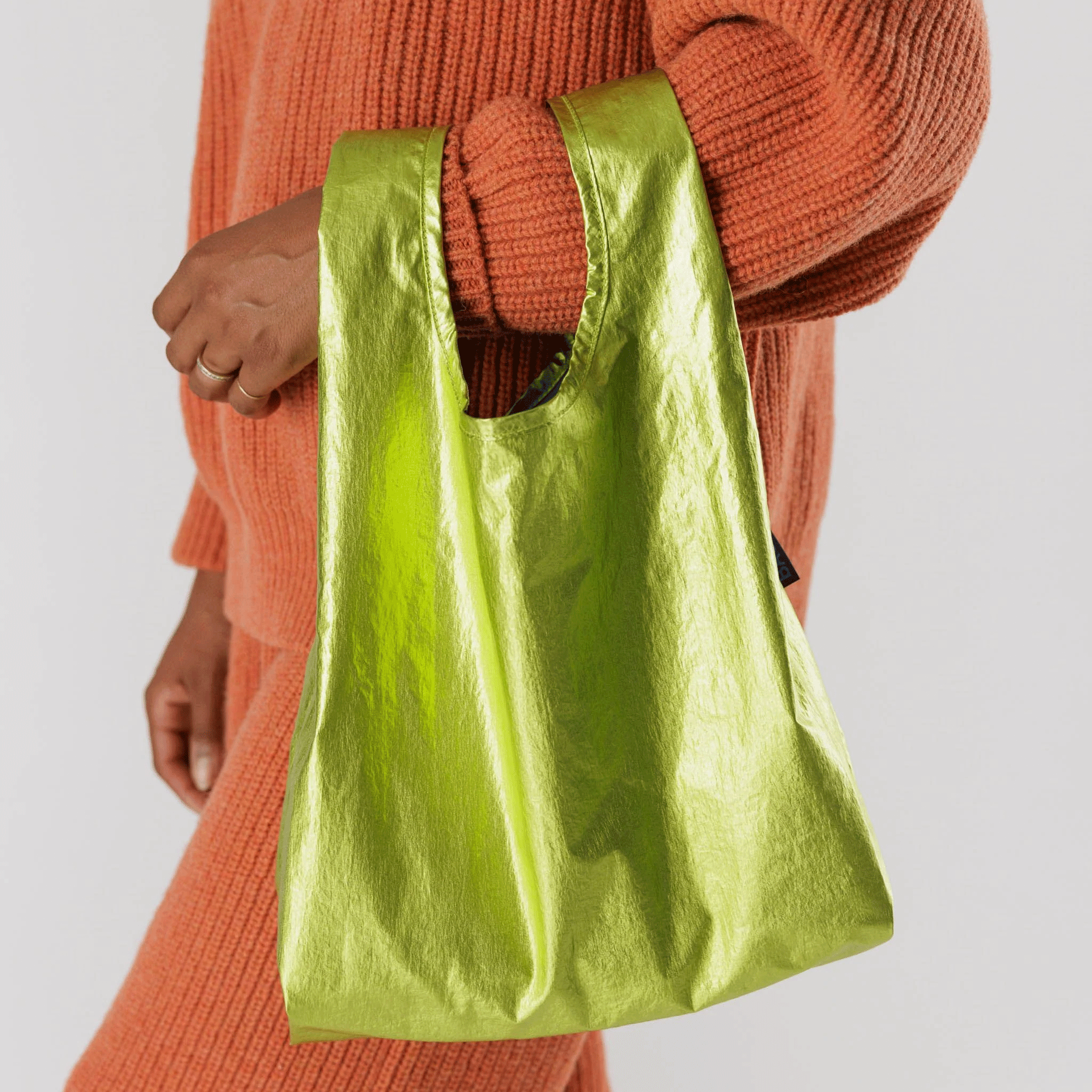 On a cream background is a model holding a neon green metallic nylon bag.