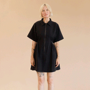 On a tan background is a model wearing a black zip up dress with short sleeves and a collar.
