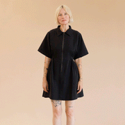 On a tan background is a model wearing a black zip up dress with short sleeves and a collar.