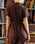 A model standing in front of a bookcase with a chocolate brown utility jumpsuit on with a zipper going up the front, a collar and a cinched waist.