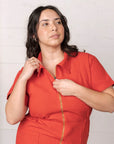 On a white background is a model wearing a short sleeve jumpsuit with a collar, side pockets and a gold zipper going down the front.