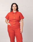 On a white background is a model wearing a short sleeve jumpsuit with a collar, side pockets and a gold zipper going down the front.