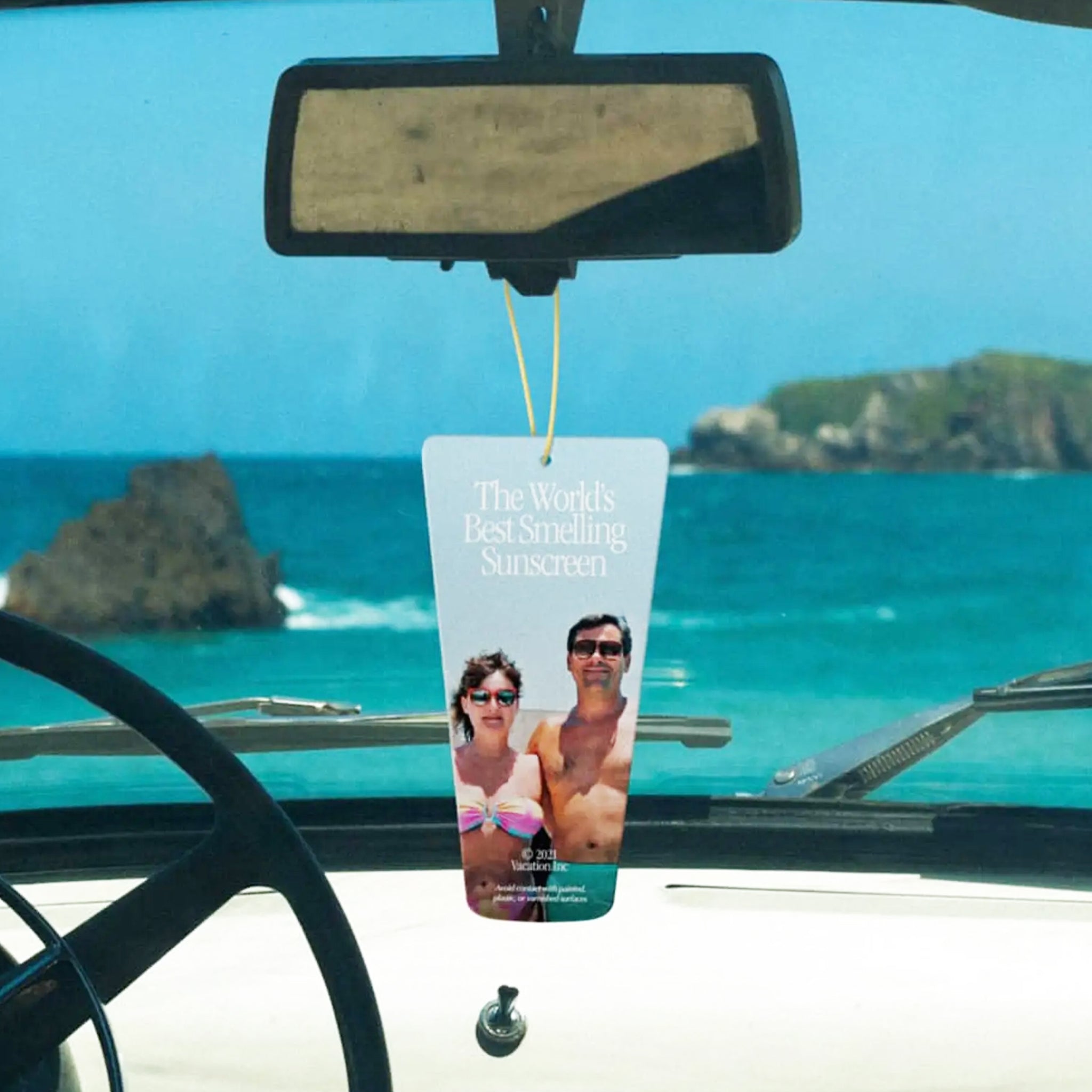 A sunscreen bottle shaped air freshener on a yellow elastic loop for hanging with a photo of a couple in swimsuits standing on a beach and text above that reads, &quot;The World&#39;s Best Smelling Sunscreen&quot;. 