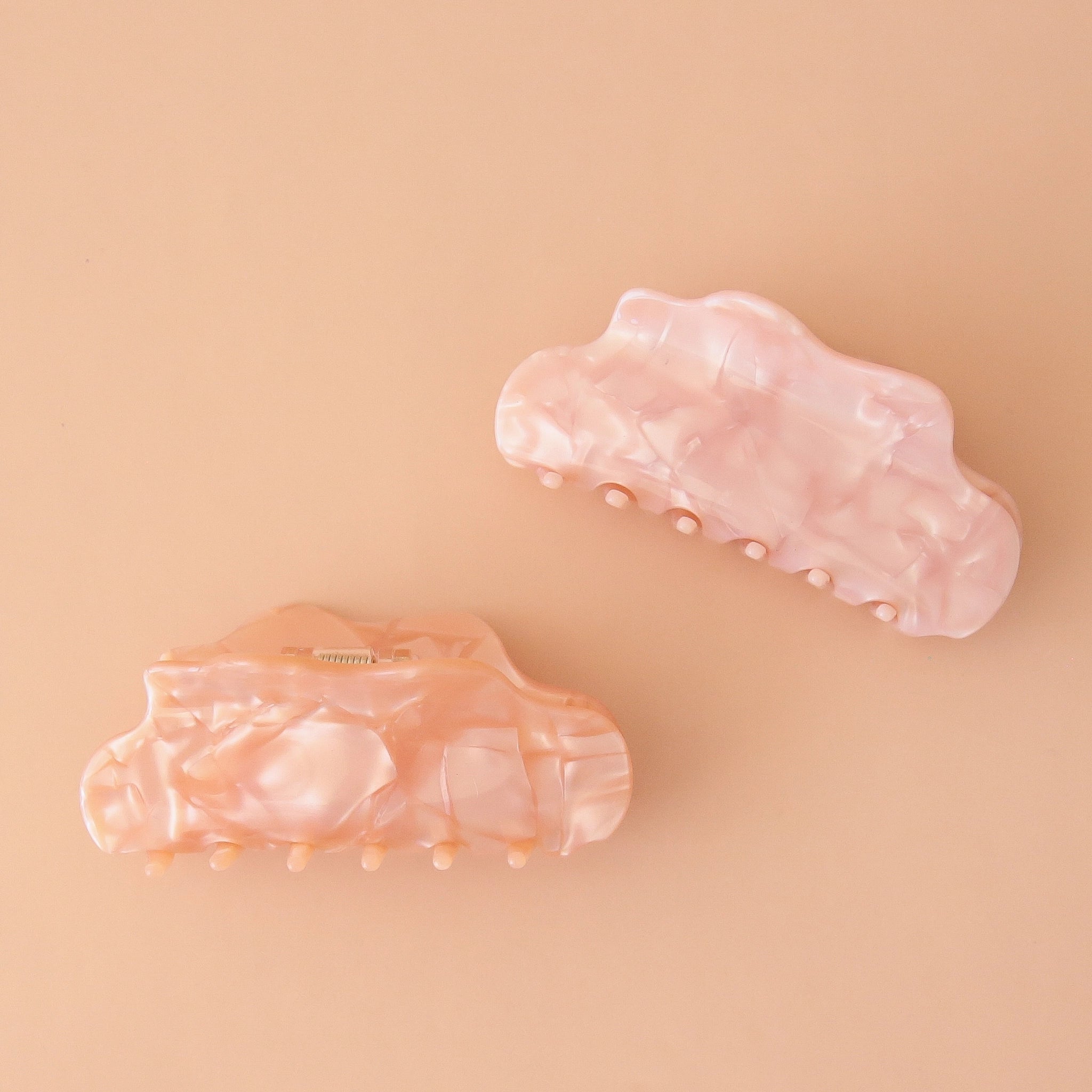 On a light pink background is the venus hair claw clip in the shade salt pink which is a light pink color with a shiny marbled finish photographed with the shade peach shell.