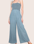 A light blue denim smocked jumpsuit with tie shoulder straps.