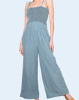 A light blue denim smocked jumpsuit with tie shoulder straps.