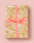 Gold wrapping paper with white leaf and flower designs sits on a pink background.