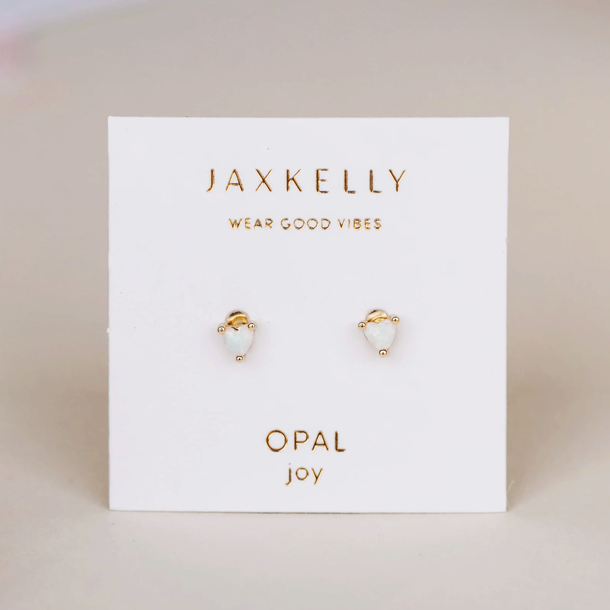 On a tan background is a pair of tiny heart shaped stud earrings with a white opal center. 