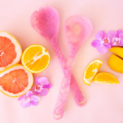 Pink salad servers shown against a pink background with sliced oranges and grapefruit, and pink orchid flowers.