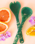 Dark green salad servers shown against a pink background with sliced oranges and grapefruit, and pink orchid flowers.