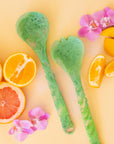 Light green salad servers shown against a yellow background with sliced oranges and grapefruit, and pink orchid flowers.