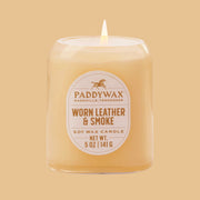 On an orange background is a neutral glass jar candle with an oval label that reads, "Paddywax Worn Leather & Smoke".
