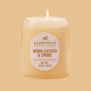 On an orange background is a neutral glass jar candle with an oval label that reads, "Paddywax Worn Leather & Smoke".