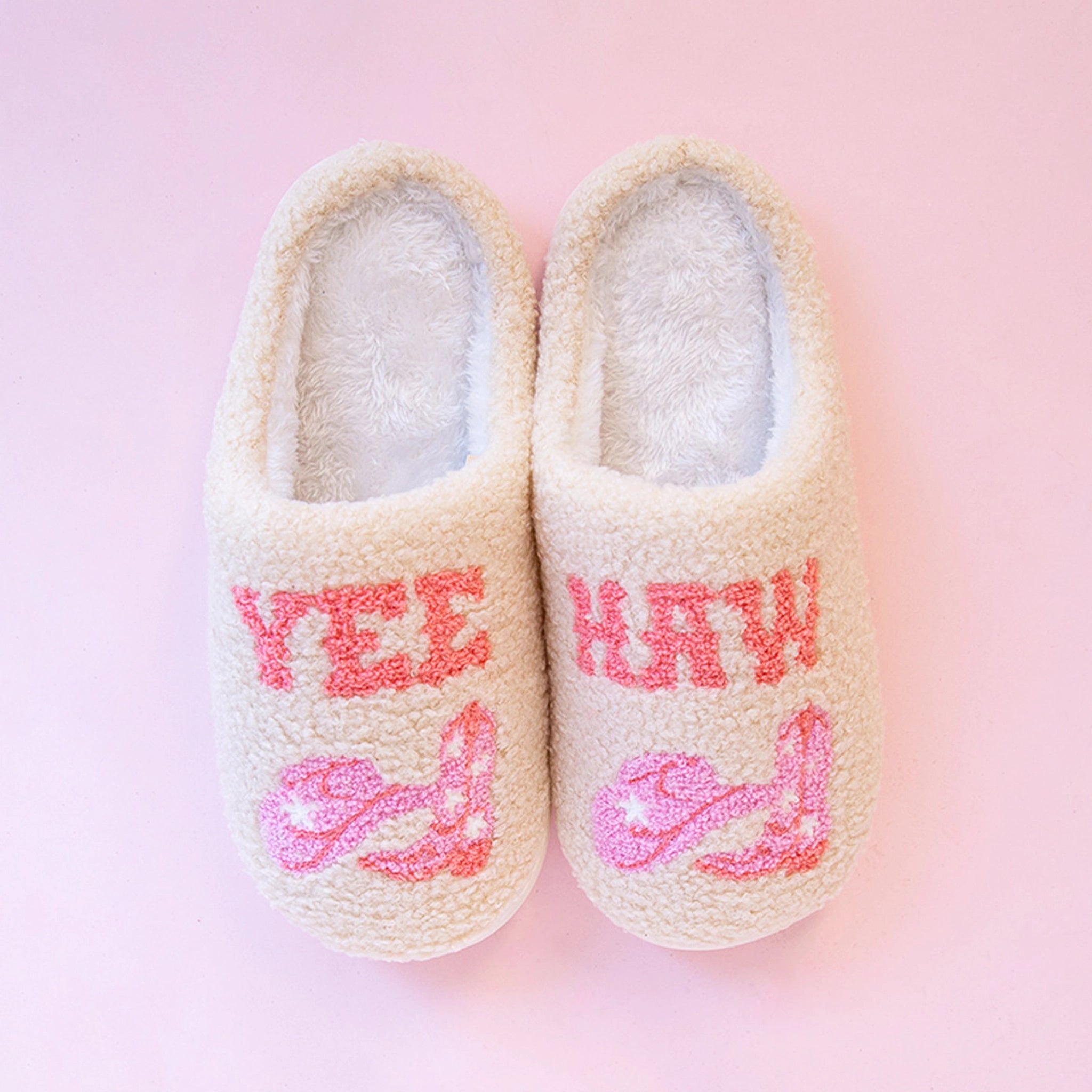 On a pink background is a pair of ivory slippers with pink western text that reads, &quot;Yee&quot; on one slipper and &quot;Haw&quot; on the other along with a graphic of pink cowgirl hats and cowgirl boots. 