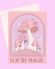 On a light purple background is a pink and purple card with an arch and mushroom design as well as text along the bottom that reads, "You're Magic". A white envelope is included with purchase, also shown here.