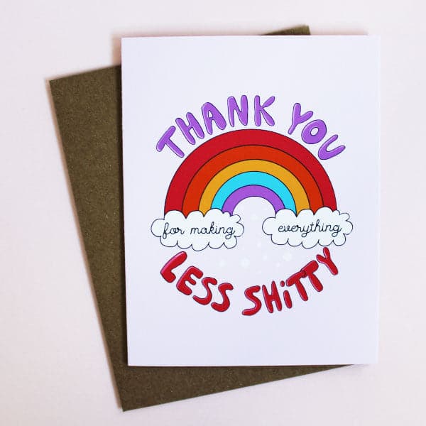 A white card with a rainbow graphic along with text above, below and inside the clouds underneath the ends of the rainbow that read, &quot;Thank you for making everything less shitty&quot; along with a brown envelope.