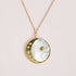 Hanging in front of a soft pink background is a gold chain necklace. At the end of the chain is a round pendent. The pendent is pearl with a small gold star in the middle. On the left side gold crescent moon with etched in stars and diamonds. 